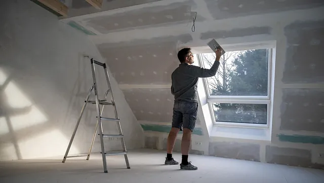 Mann pusser og sparkler gipsvegger i lyst rom med takvindu og stige